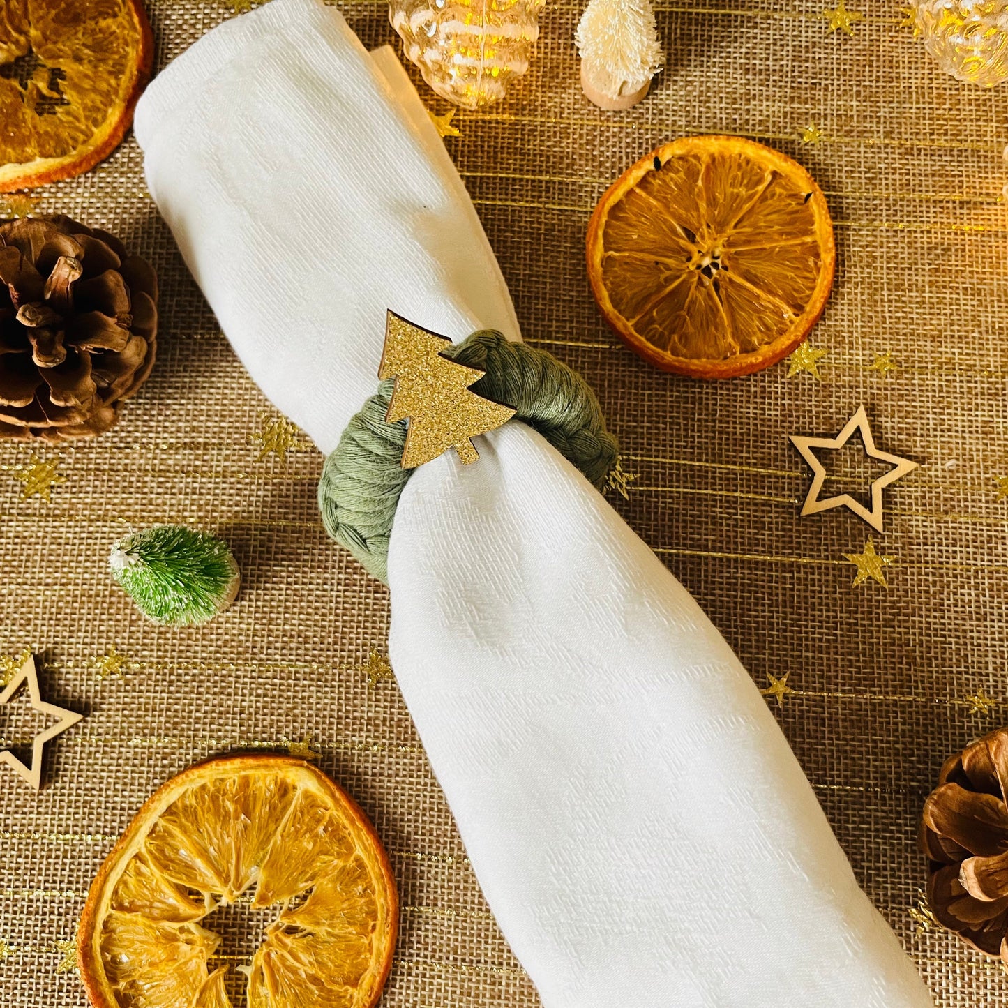 White napkin with a green macrame wreath napkin ring with a gold tree embellishment on a textured tablecloth with Christmas decorations.