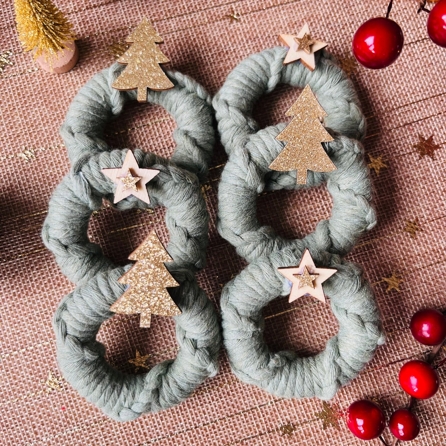 Green macrame wreath napkin rings with gold tree and star decorations on a burlap surface with dried oranges and pine cones.