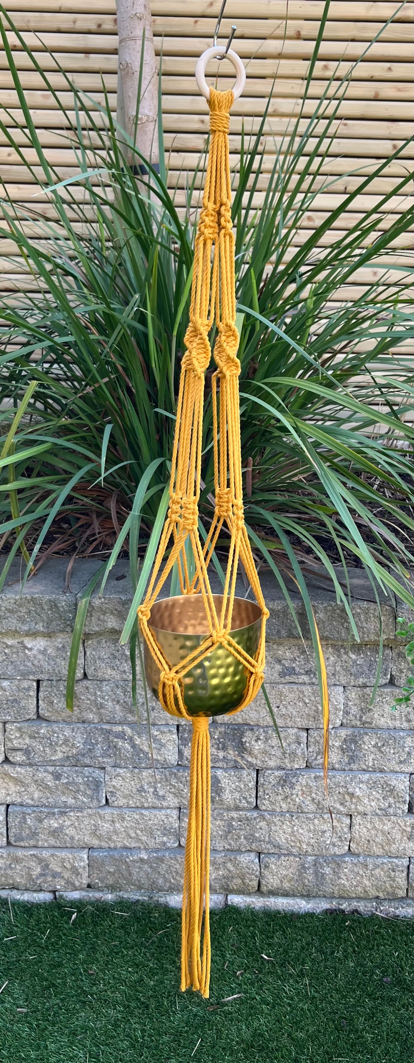 Macramé plant hanger in mustard with a gold pot against a garden background