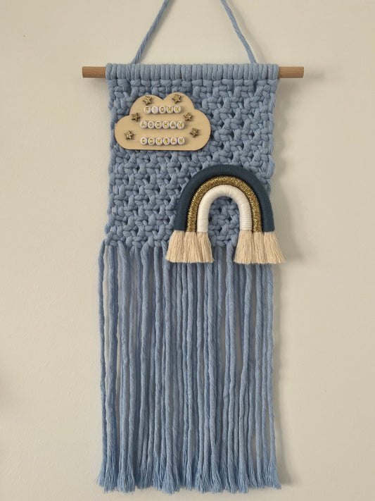 Blue macramé wall hanging with cloud and rainbow design on a white background