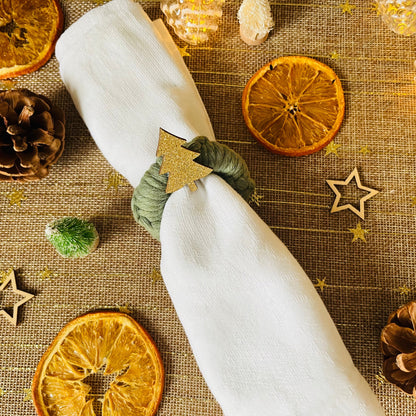 White napkin with a green macrame wreath napkin ring with a gold tree embellishment on a textured tablecloth with Christmas decorations.