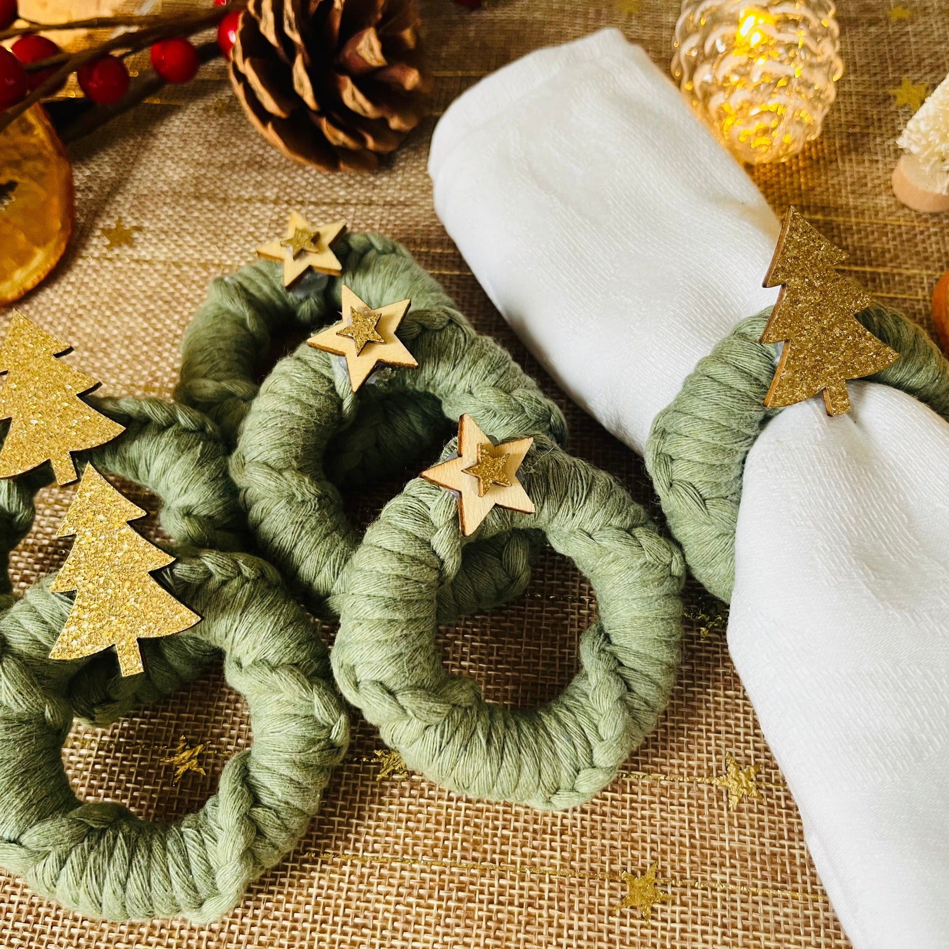 Green macrame wreath napkin rings with gold tree and star decorations on a burlap surface with dried oranges and pine cones.