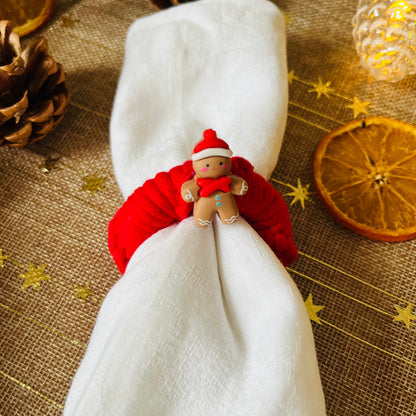White napkin with a red macrame wreath napkin ring with a festive embellishment on a textured tablecloth with Christmas decorations.