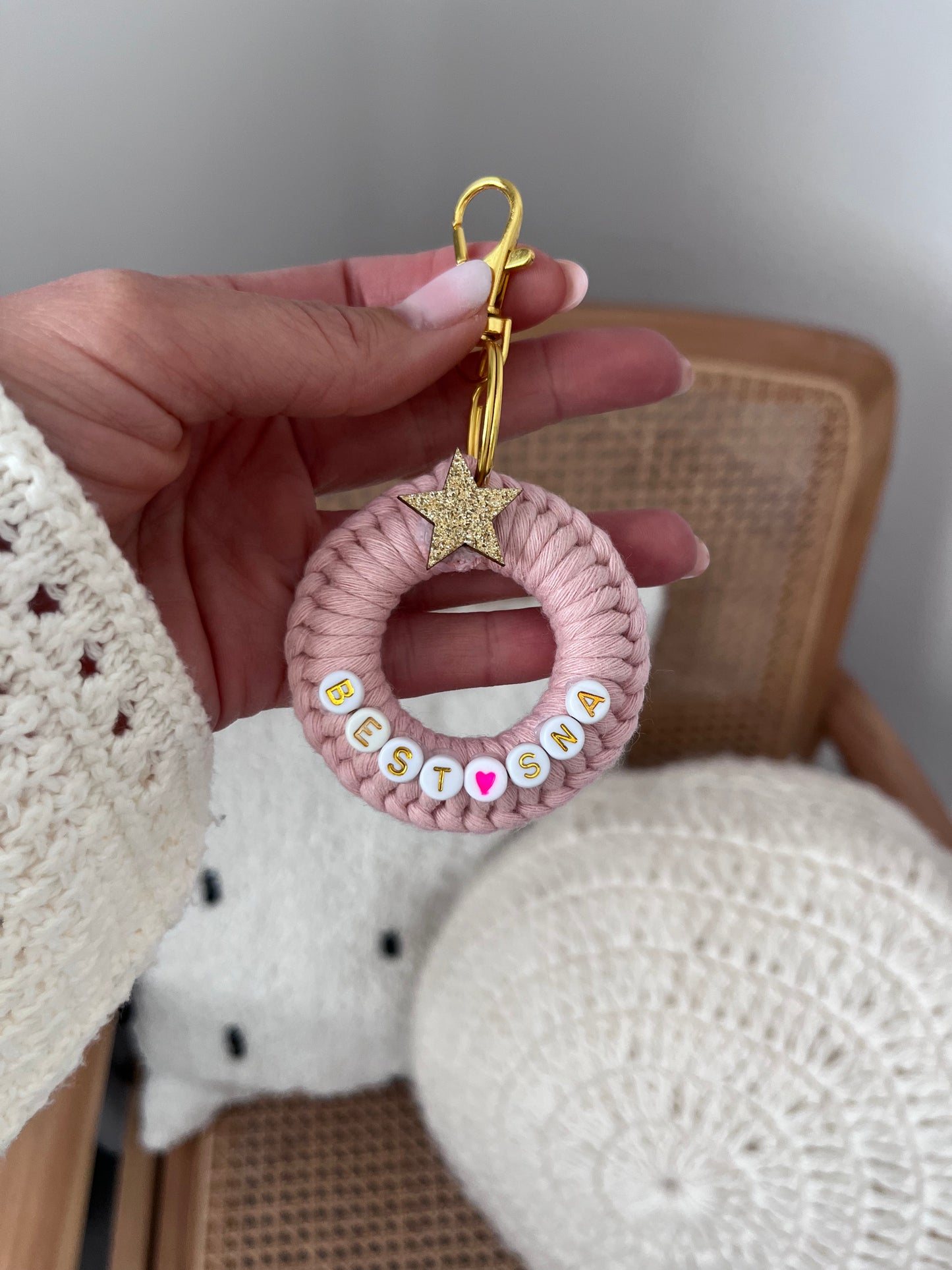 Pink crocheted keychain with letters and a gold star held by a hand against a neutral background.