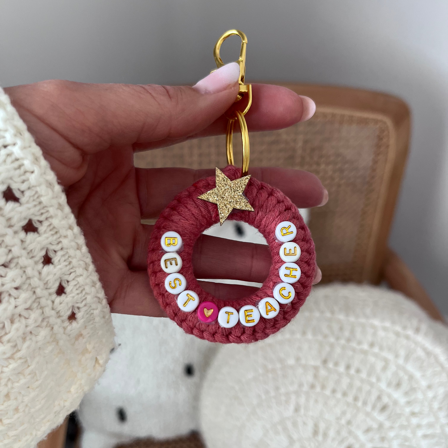 Hand holding a pink crocheted keychain with white letters and a gold star, with a neutral background.