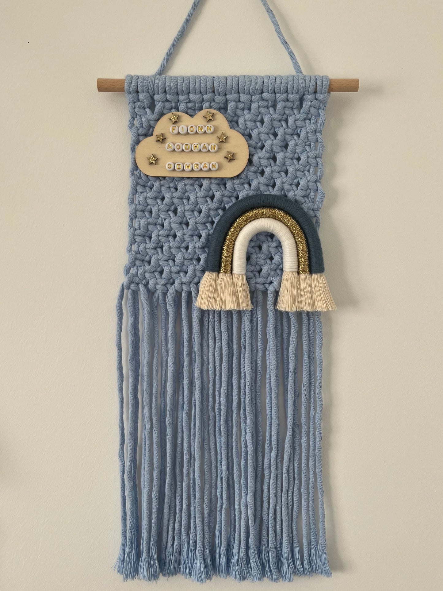 Blue macramé wall hanging with cloud and rainbow design on a white background