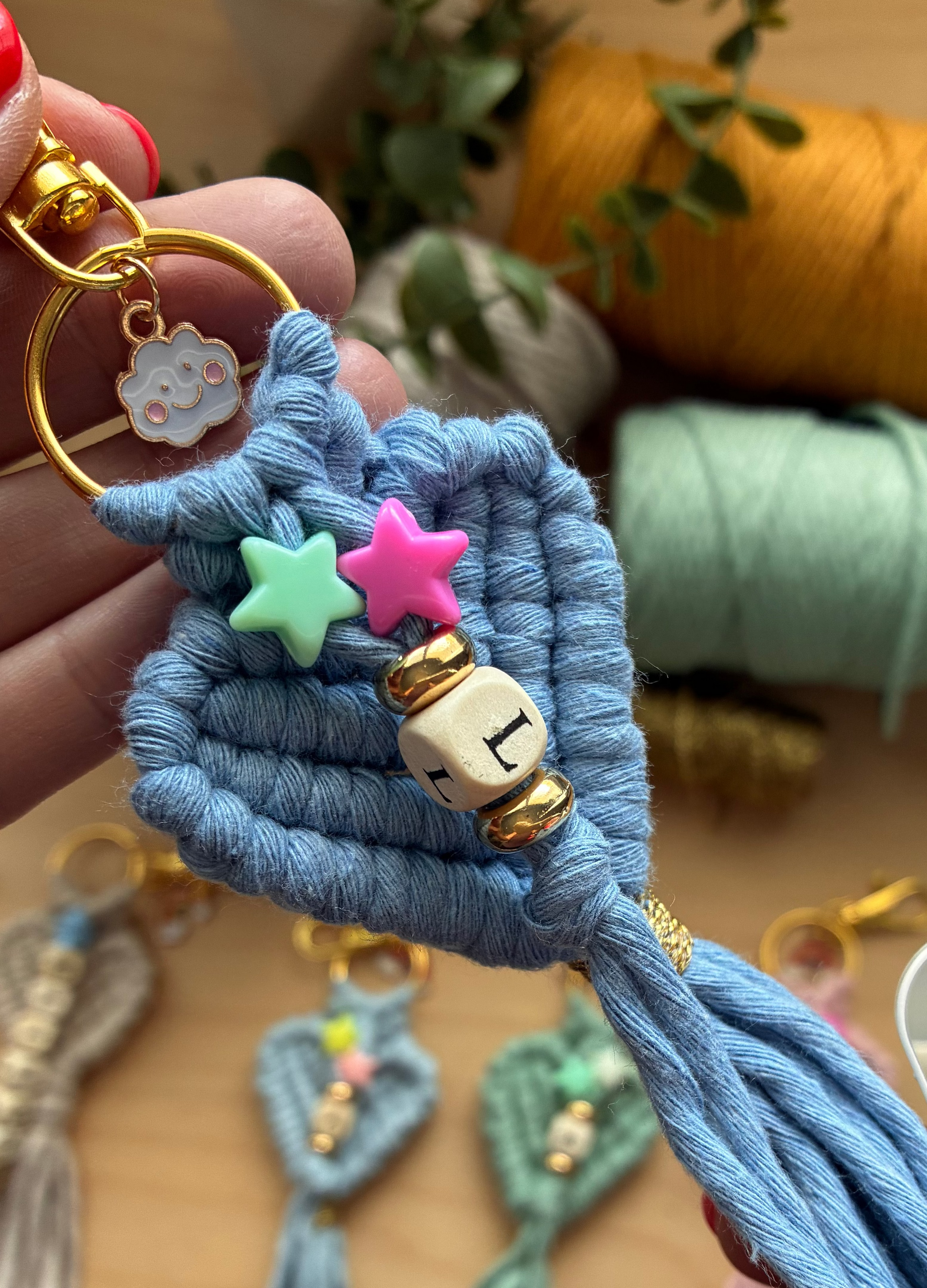 A handmade macrame keychain held by a hand, featuring a light blue knotted cord heart with a gold key ring, a white and gold beaded charm, and pastel star accents. Other macrame keychains are blurred in the background on a wooden surface.