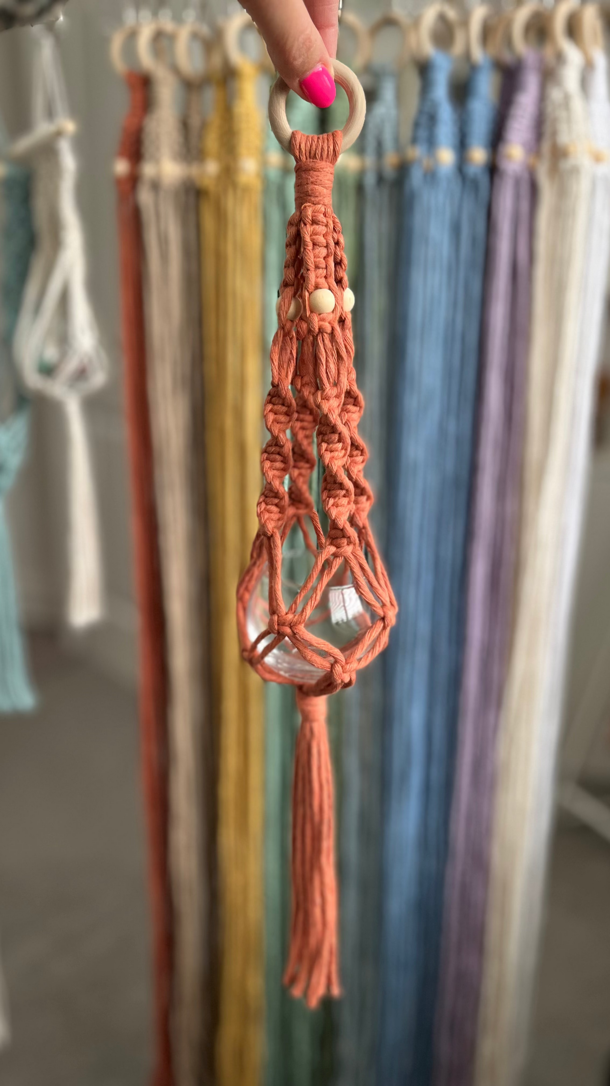 Terracotta macrame crystal hanger with a  wooden ring held by a hand against a colorful fabric background.