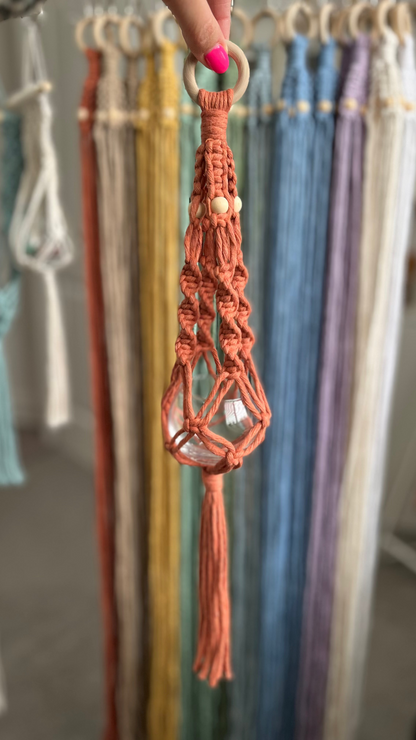 Terracotta macrame crystal hanger with a  wooden ring held by a hand against a colorful fabric background.